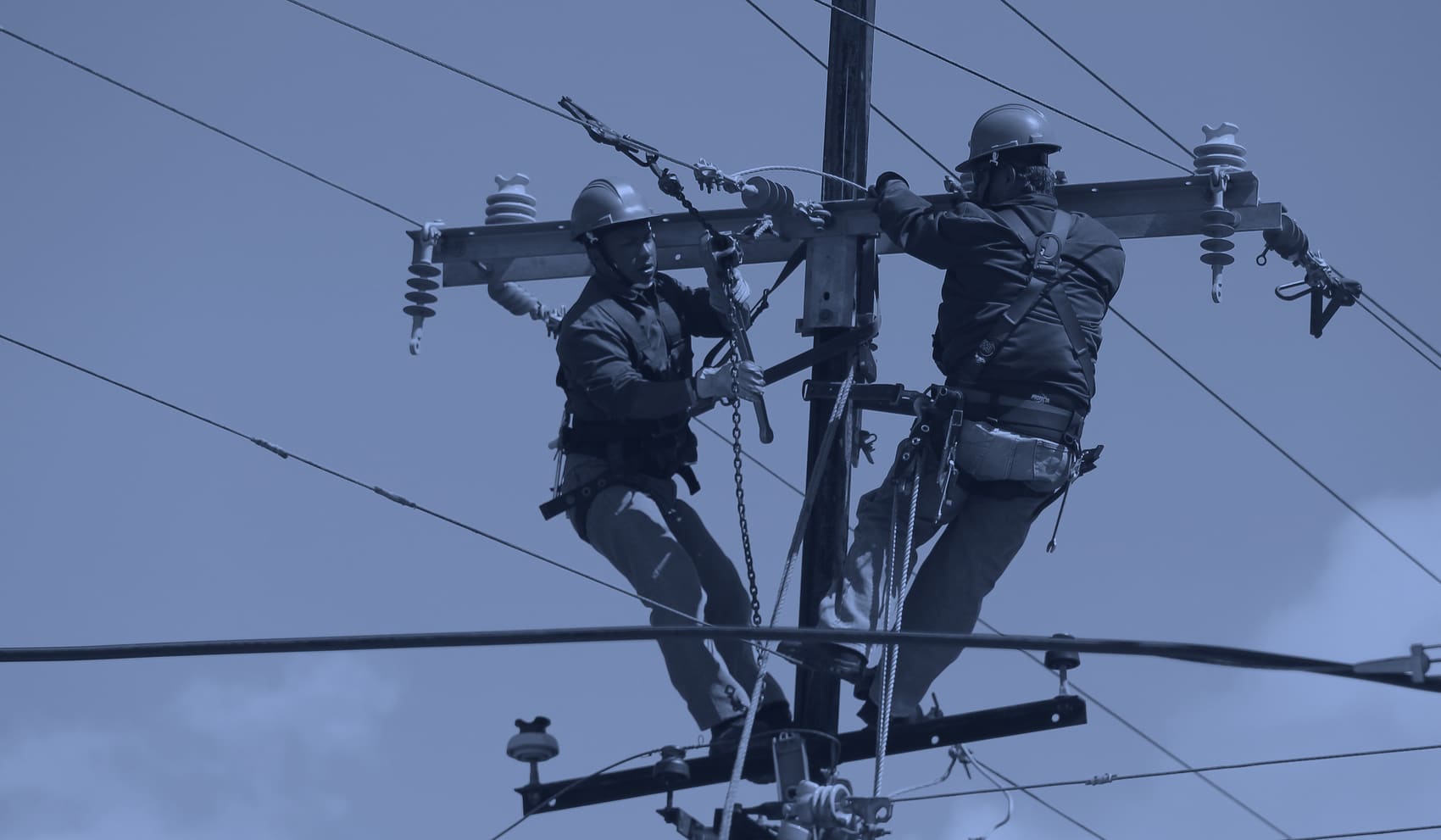 Two line workers in fall-protection harnesses making up a crossarm at the top of a distribution pole